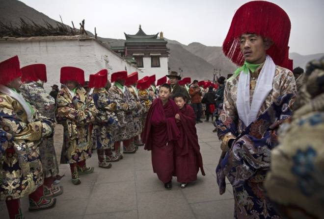 Young monks walk past men in traditional costumes preparing for a procession.